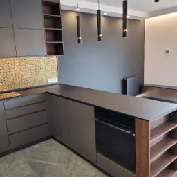 Nowoczesna kuchnia z szarymi szafkami, blatem z ciemnego kamienia, złotą mozaiką nad blatem i wiszącymi cylindrycznymi lampami. Na podłodze marmurkowe płytki w odcieniach szarości.