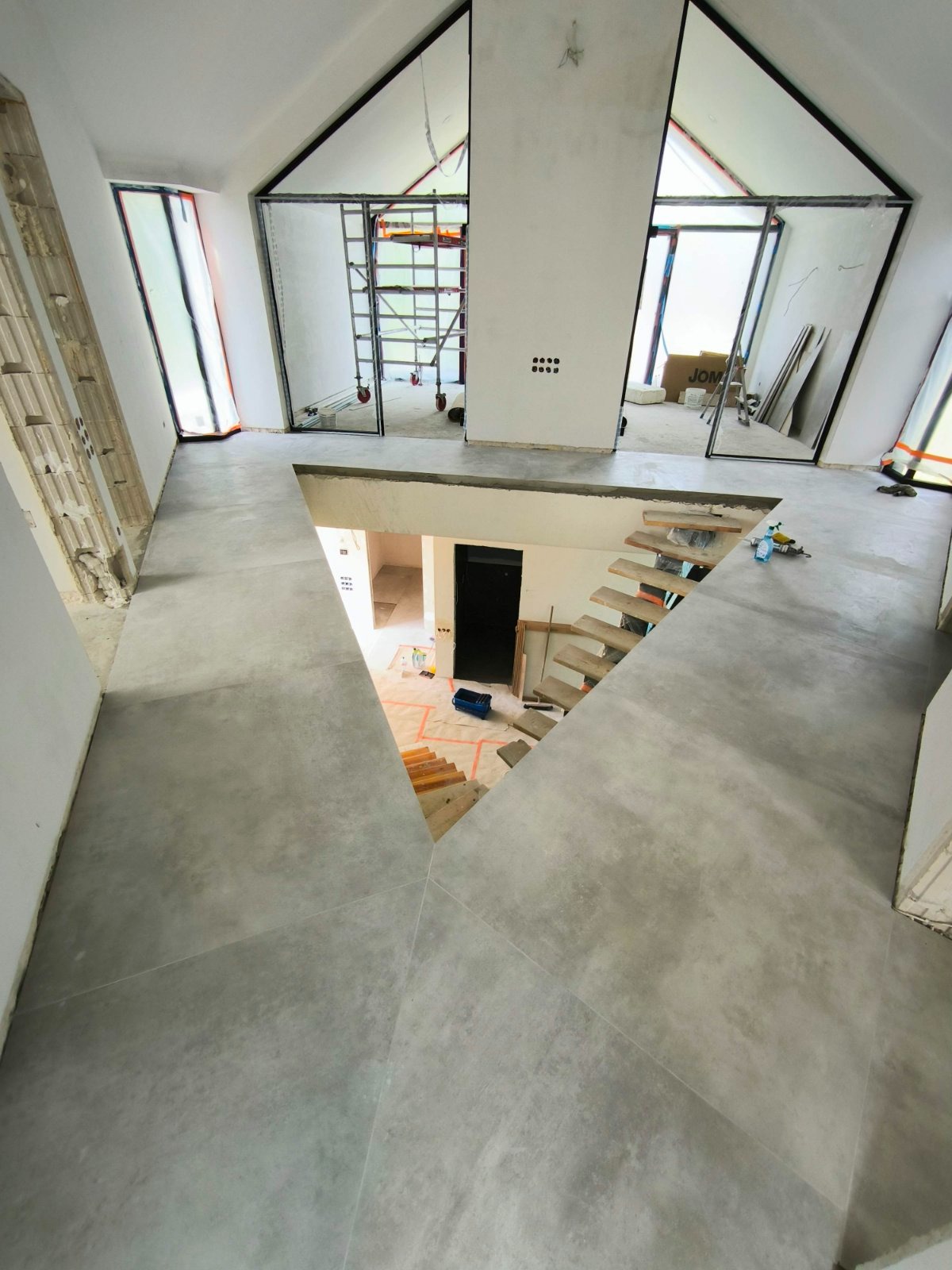 Wnętrze domu w trakcie remontu: szara wylewka na podłodze, surowe drewniane schody, widoczne rusztowania i okna z czarnymi ramami, częściowo zabezpieczone folią. Na podłodze narzędzia i materiały...