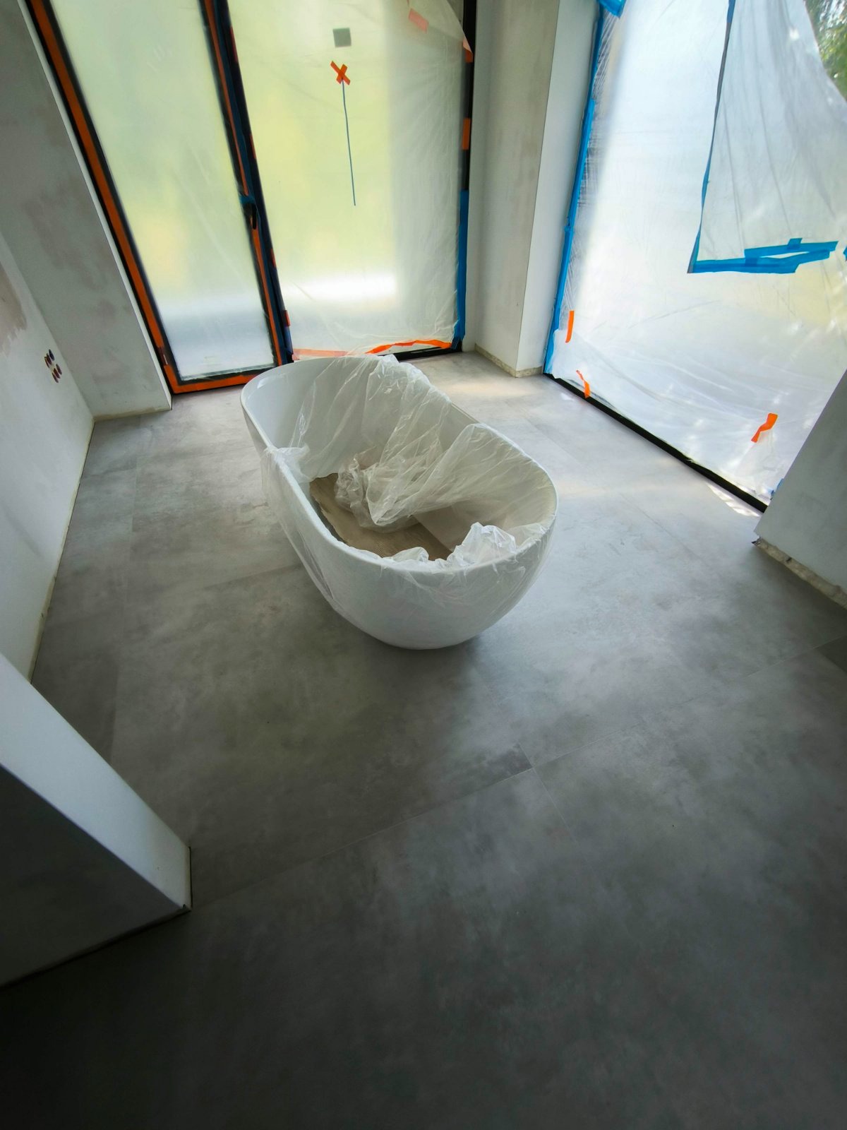 Wanna owalna zabezpieczona folią w trakcie prac wykończeniowych, okna i drzwi również zabezpieczone folią malarską, szara posadzka z płytek.