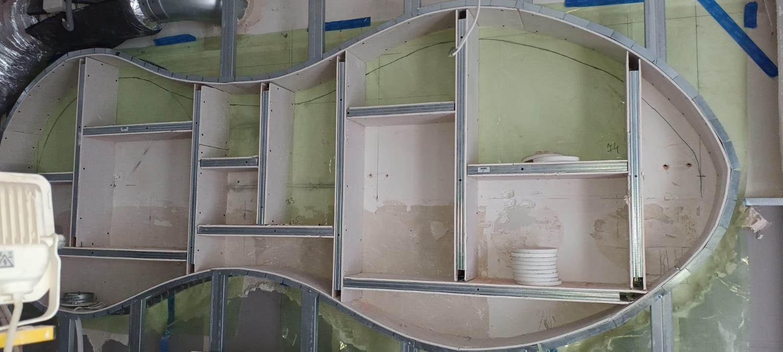 Nietypowa konstrukcja szkieletowa z profili metalowych i płyt gipsowo-kartonowych w kształcie nerki, tworząca wnęki na ścianie, widoczne ślady po ołówku i wstępne szpachlowanie.