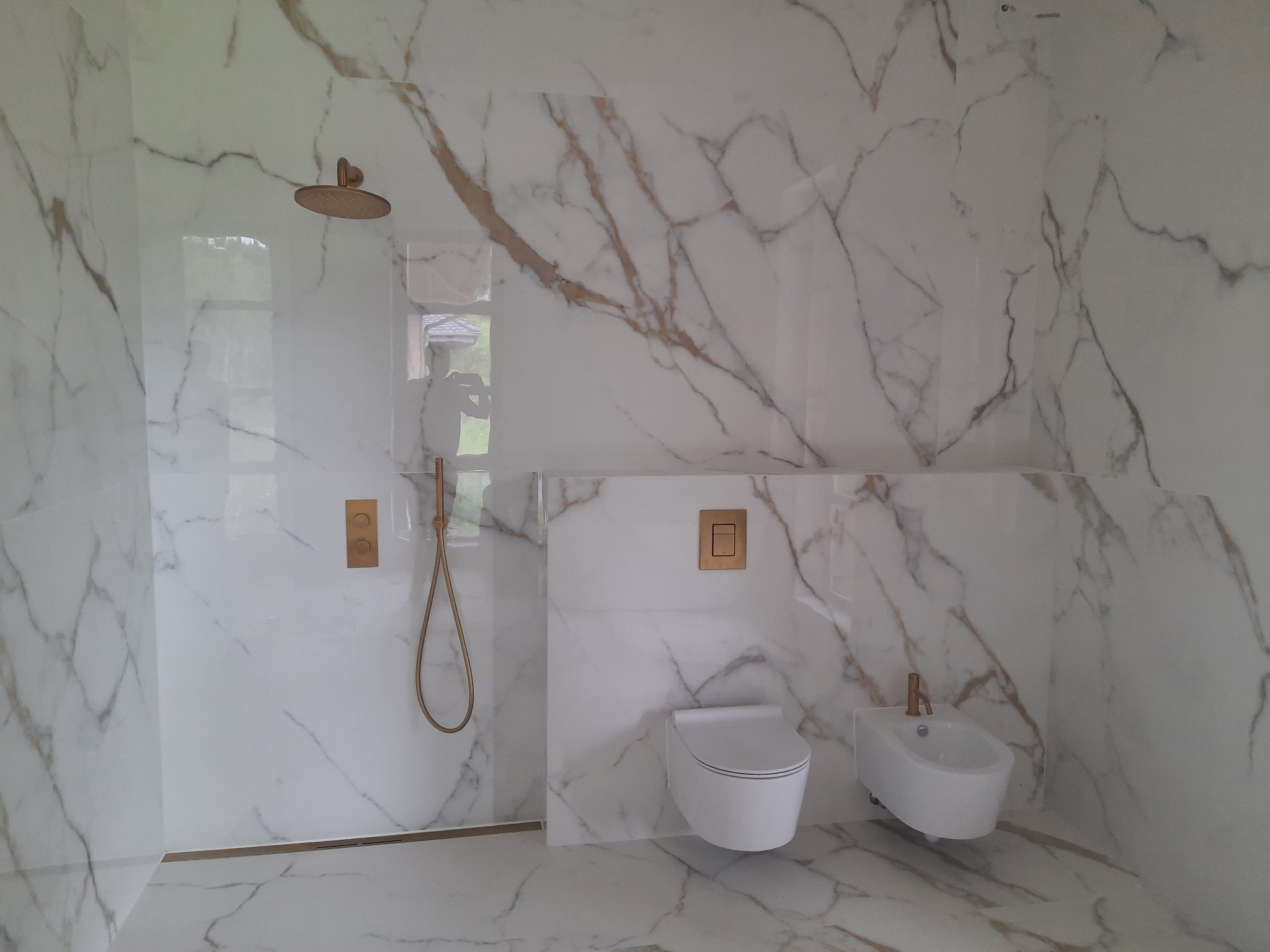 Wykończona łazienka w Halinowie z białymi płytkami imitującymi marmur z brązowymi żyłami, złotą armaturą, wiszącą toaletą i bidetem.