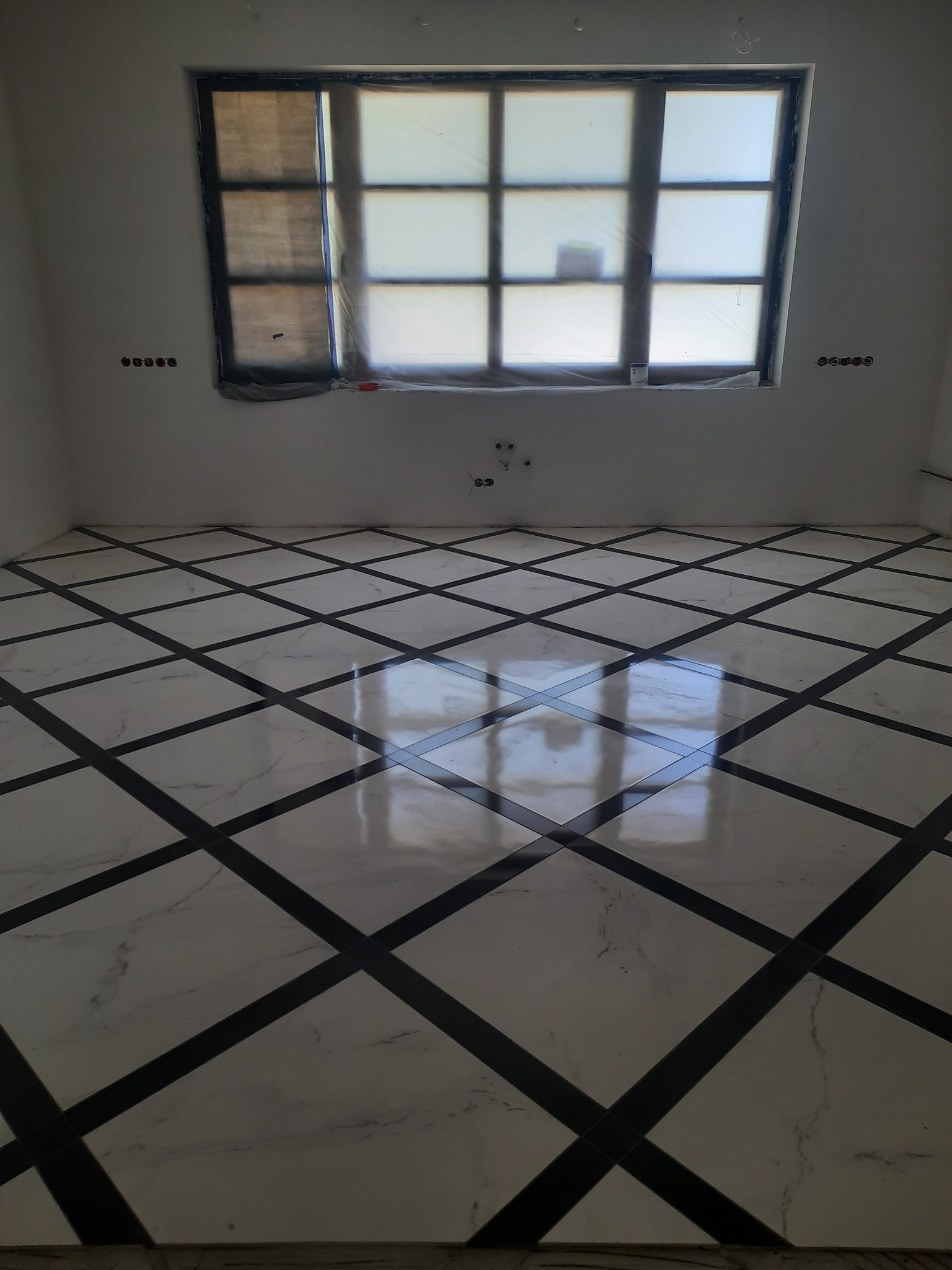 Nowoczesna podłoga z białych płytek marmurowych z czarnymi wstawkami, ułożonych w karo, w jasnym pomieszczeniu z oknem zabezpieczonym folią malarską.