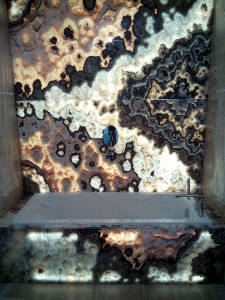 Podświetlana wanna z obudową i ścianą z onyksu o nieregularnym wzorze w odcieniach brązu, bieli i szarości, bateria wannowa na boku wanny