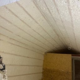 P.H.U.AKRO Marek Zalewski - Ocieplanie poddasza pianką PUR w Legionowie. Widoczne drewniane elementy konstrukcji i nałożona warstwa izolacyjna. Praca w trakcie wykonywania, osoba w kombinezonie ochronnym.