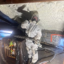 P.H.U.AKRO Marek Zalewski - Pracownik w kombinezonie ochronnym aplikuje piankę PUR na poddaszu, widoczna struktura pianki i oświetlenie robocze. Zabezpieczone otoczenie.