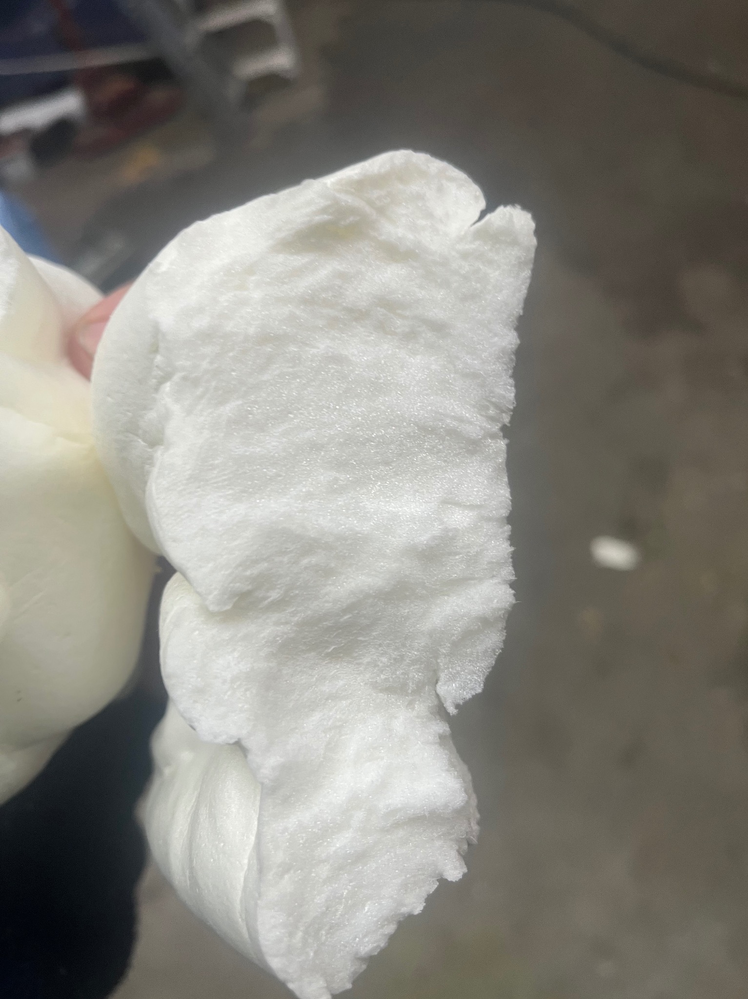 Fragment białej, spienionej pianki PUR o nieregularnej strukturze, trzymanej w dłoni, prezentowany w zbliżeniu na tle szarej powierzchni.