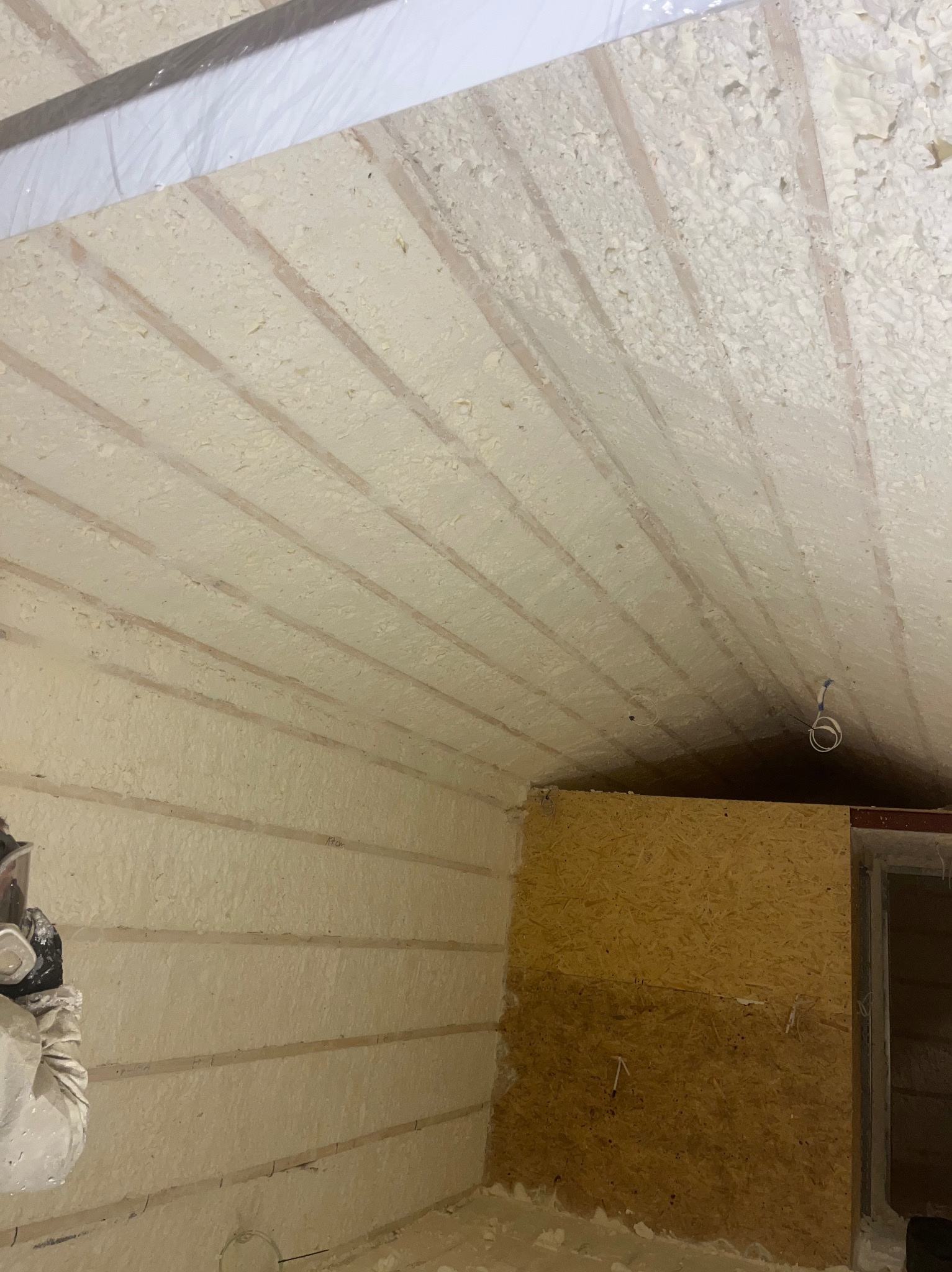 Ocieplanie poddasza pianką PUR w Legionowie. Widoczne drewniane elementy konstrukcji i nałożona warstwa izolacyjna. Praca w trakcie wykonywania, osoba w kombinezonie ochronnym.