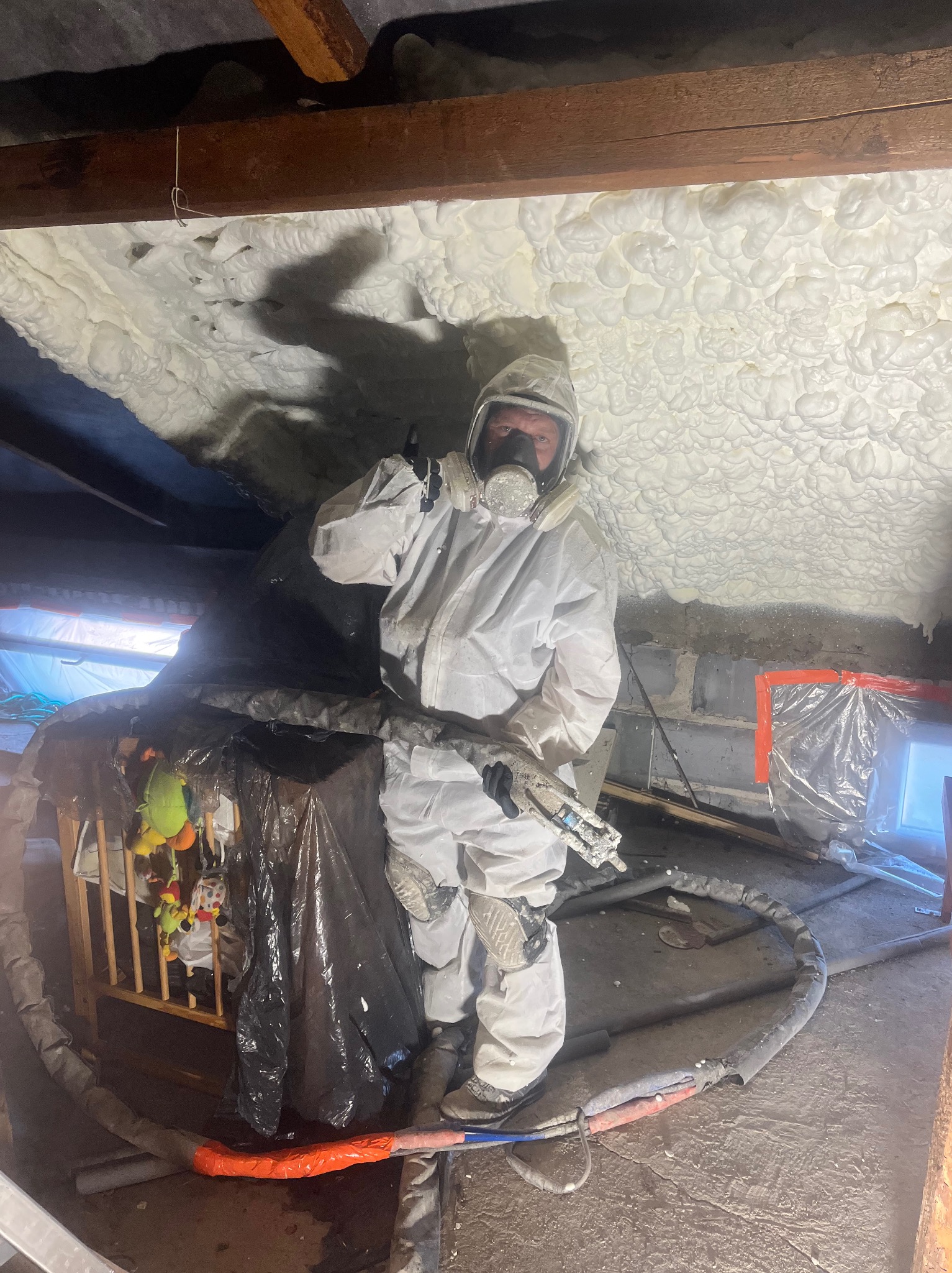 Pracownik w kombinezonie ochronnym aplikuje piankę PUR na poddaszu, widoczna struktura pianki i oświetlenie robocze. Zabezpieczone otoczenie.