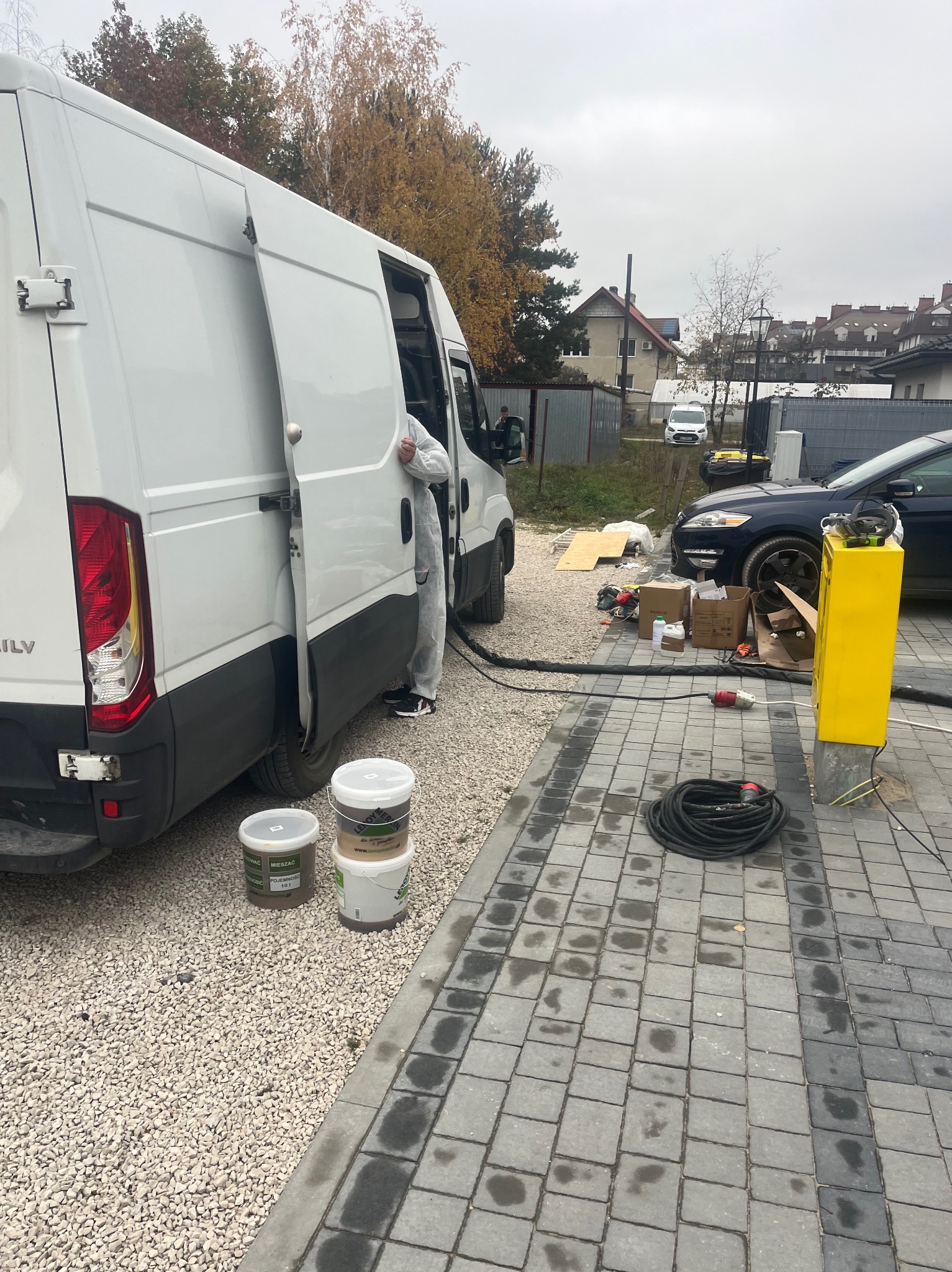 Pracownik w kombinezonie przy otwartym busie z narzędziami i pojemnikami. Przygotowanie do pracy na zewnątrz budynku. Widoczne kable i sprzęt.