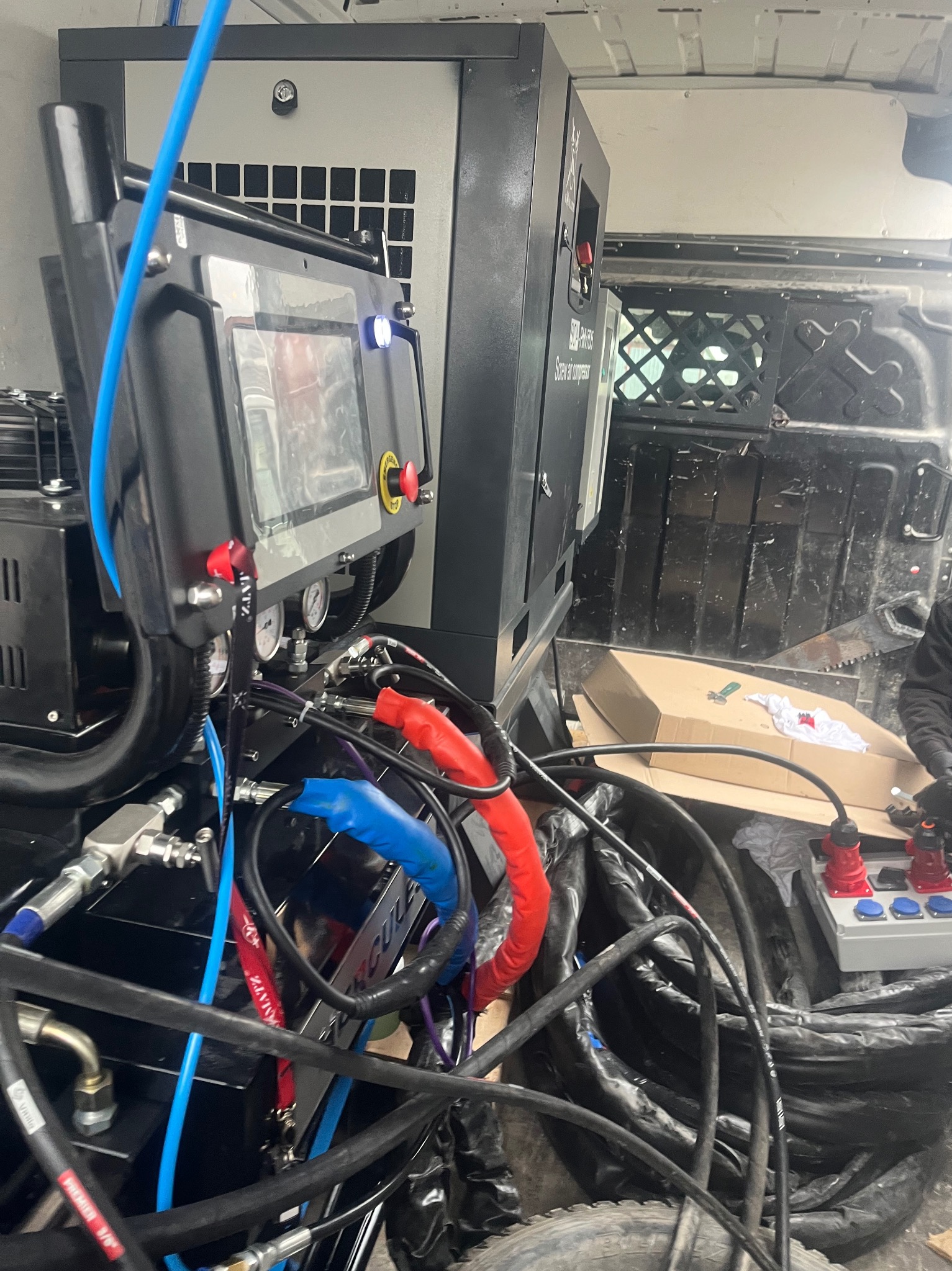 Sprzęt do natrysku pianki PUR wewnątrz furgonetki: sterownik z ekranem, kompresor, przewody hydrauliczne i zasilające. Widoczne narzędzia i akcesoria.