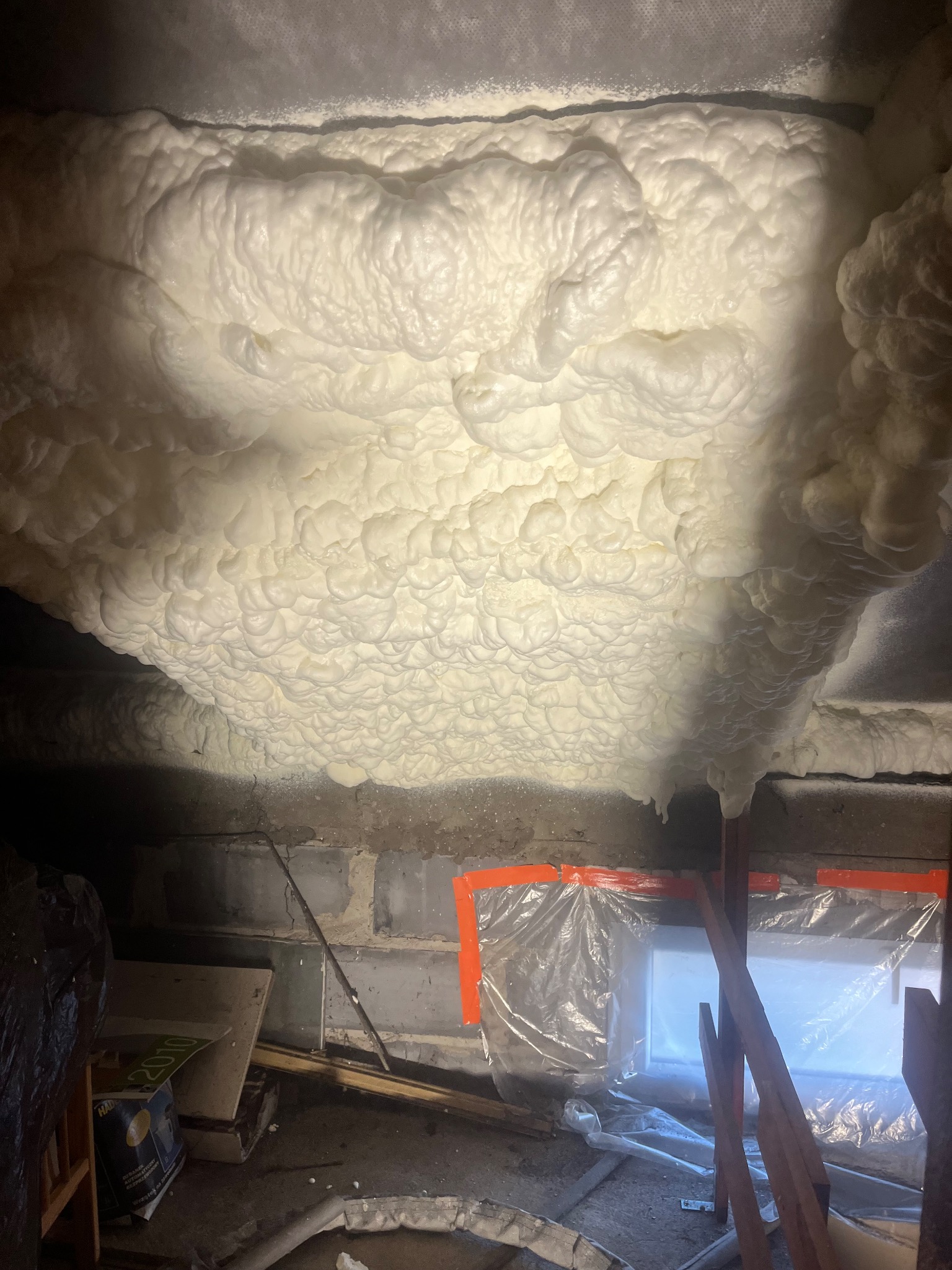 Izolacja poddasza pianką PUR: gęsta, biała piana szczelnie wypełnia przestrzeń, widoczne elementy konstrukcyjne i zabezpieczenia okienne folią.