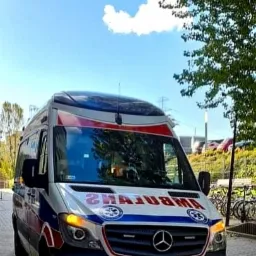 Ambulans marki Mercedes z włączonymi światłami awaryjnymi, zaparkowany na tle błękitnego nieba z pojedynczą chmurą i zieleni drzew