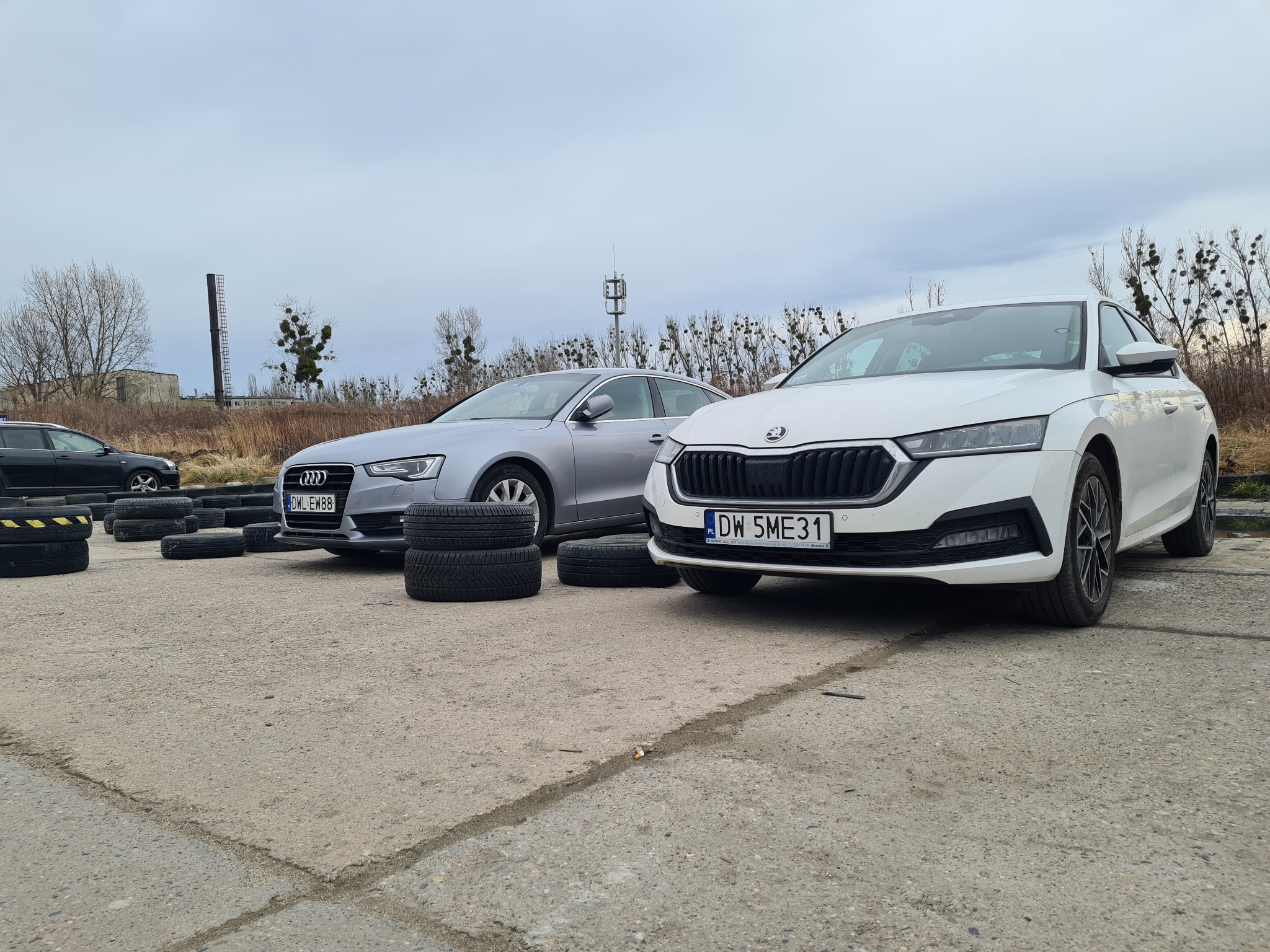 Dwa samochody osobowe (biała Skoda Octavia i srebrne Audi) stojące na betonowym placu, otoczone stosem opon. W tle widoczna wieża telekomunikacyjna i drzewa bez liści.