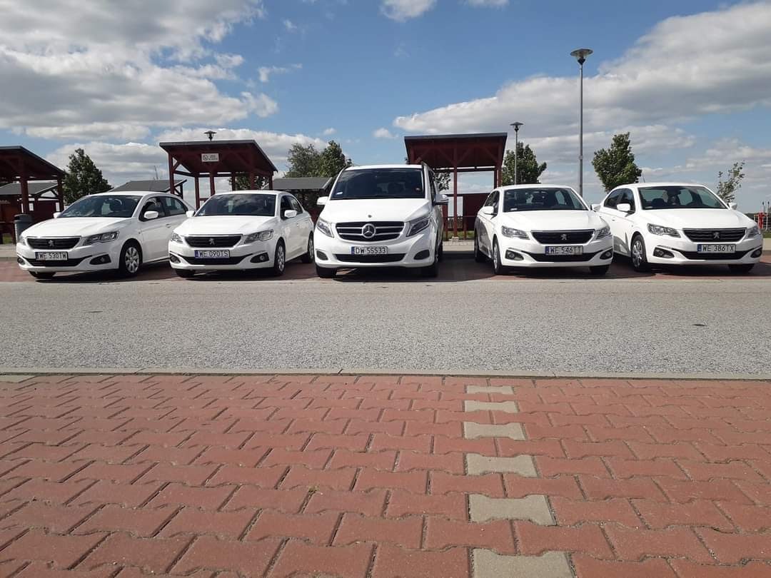 Flota pięciu białych samochodów osobowych i busa Mercedes-Benz, ustawionych w rzędzie na parkingu z kostki brukowej, błękitne niebo z chmurami w tle.