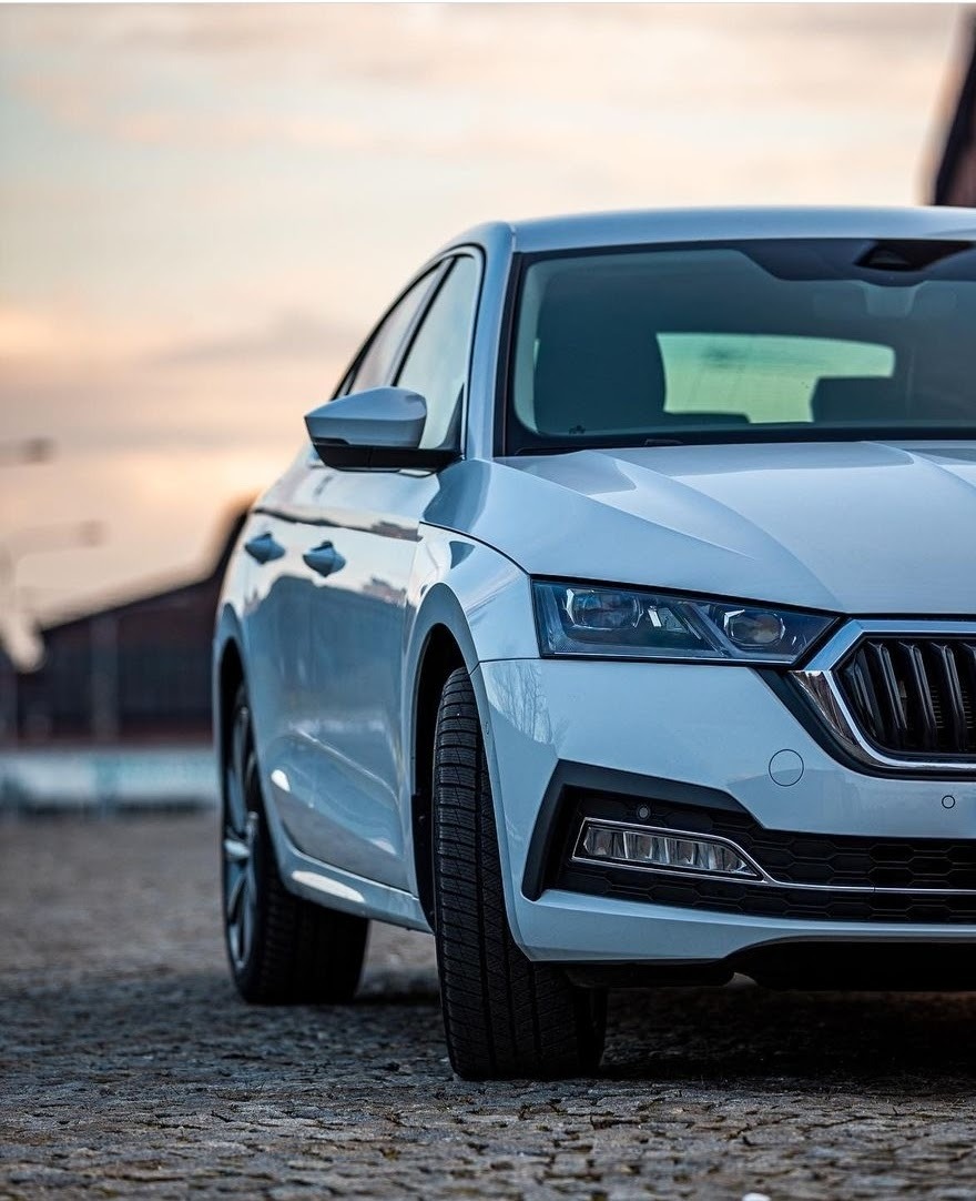 Nowoczesny, biały samochód osobowy marki Skoda Octavia, sfotografowany z niskiej perspektywy, ukazujący detale przedniego reflektora i opony na tle rozmytego zachodzącego słońca.
