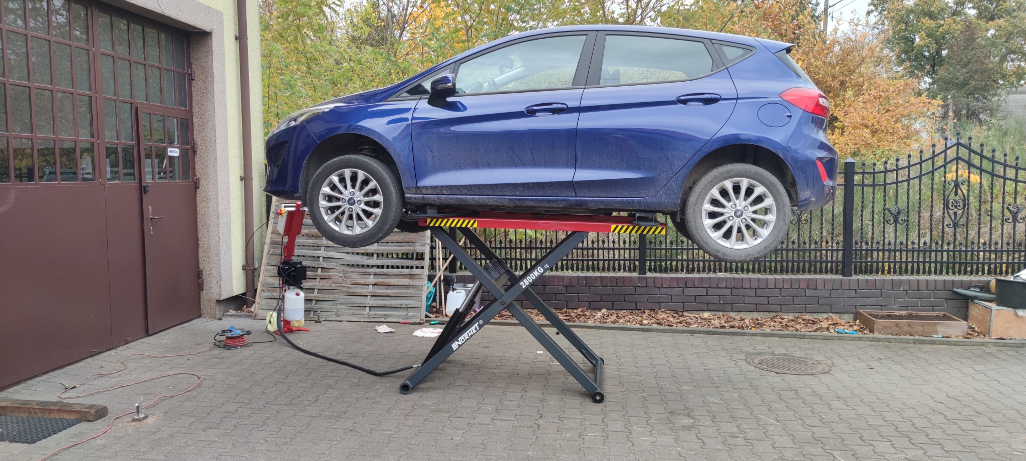 Niebieski samochód Ford Fiesta podniesiony na dwukolumnowym podnośniku warsztatowym NORMET 2800KG przed garażem z widocznym przewodem hydraulicznym i skrzynką sterującą.
