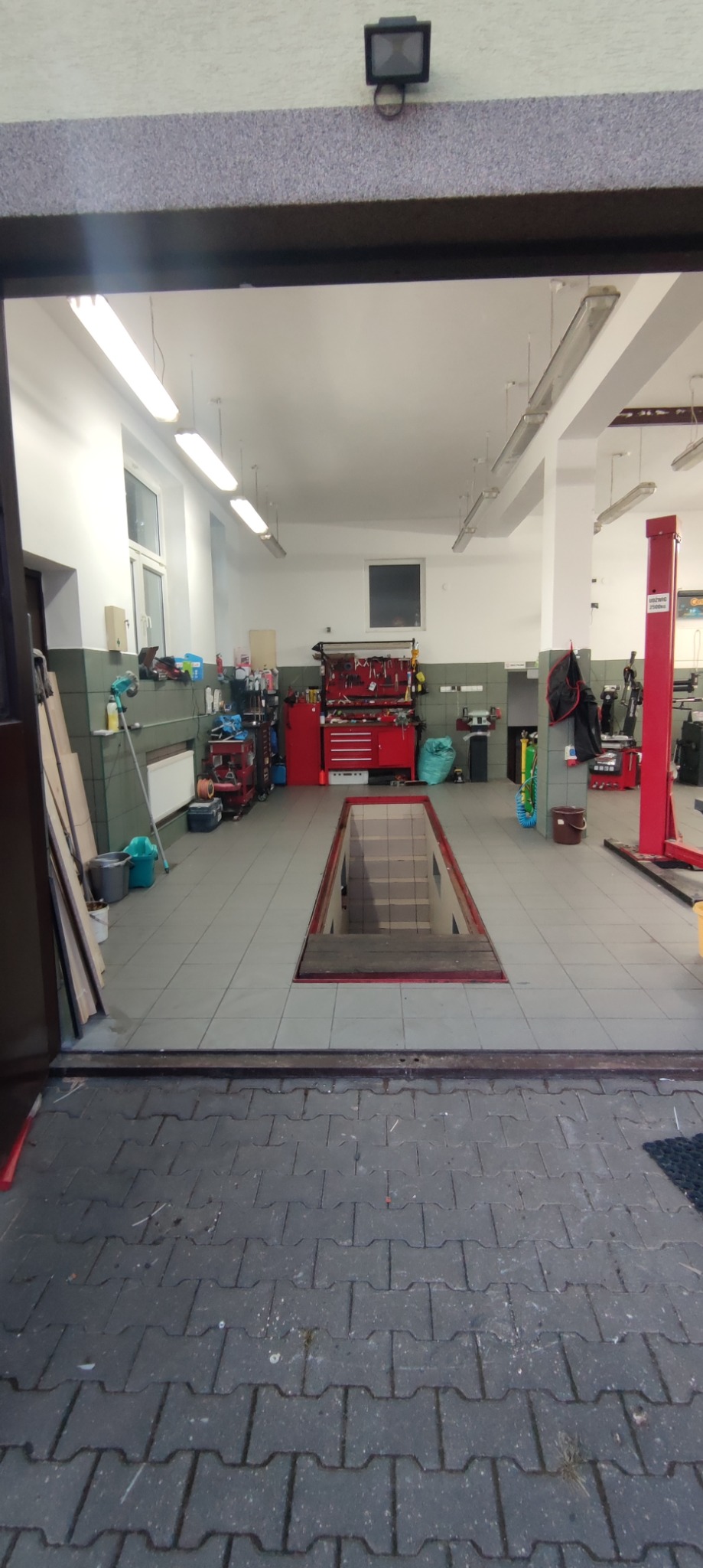 Wnętrze warsztatu samochodowego z kanałem inspekcyjnym, widok z zewnątrz, narzędzia i podnośnik widoczne w tle.