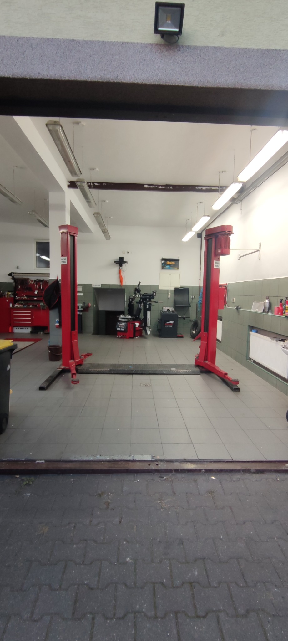 Wnętrze warsztatu samochodowego widziane z zewnątrz, z dwukolumnowym podnośnikiem w kolorze czerwonym na pierwszym planie, narzędzia i wyposażenie widoczne w tle.