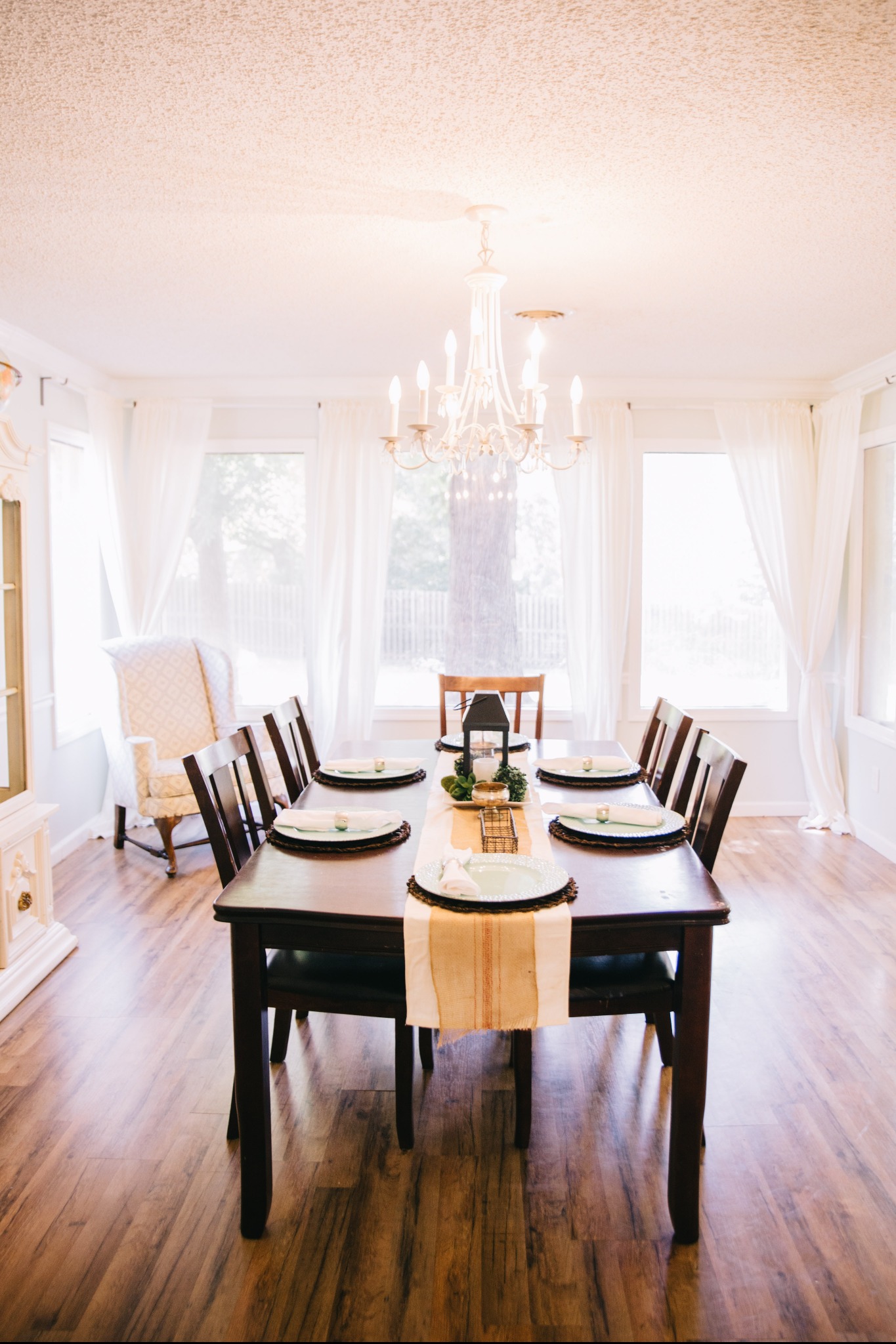 Jasne wnętrze jadalni z drewnianym stołem, nakrytym dla sześciu osób, z dekoracyjnym bieżnikiem i centralną ozdobą, oświetlone przez żyrandol w stylu vintage.