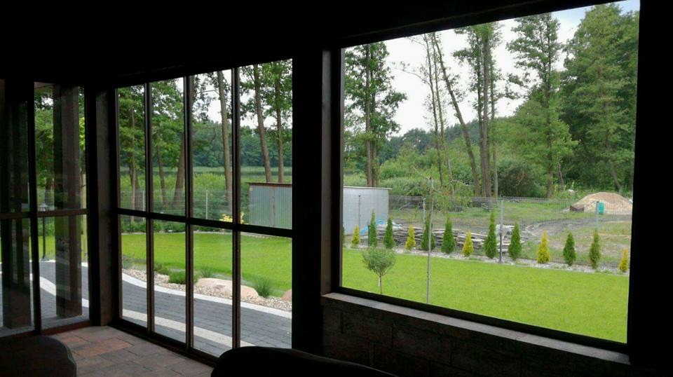 Wnętrze zadaszonego tarasu z widokiem na zadbany ogród z trawnikiem, ścieżką z kostki, rzędem małych drzewek iglastych i metalowym budynkiem gospodarczym w tle.