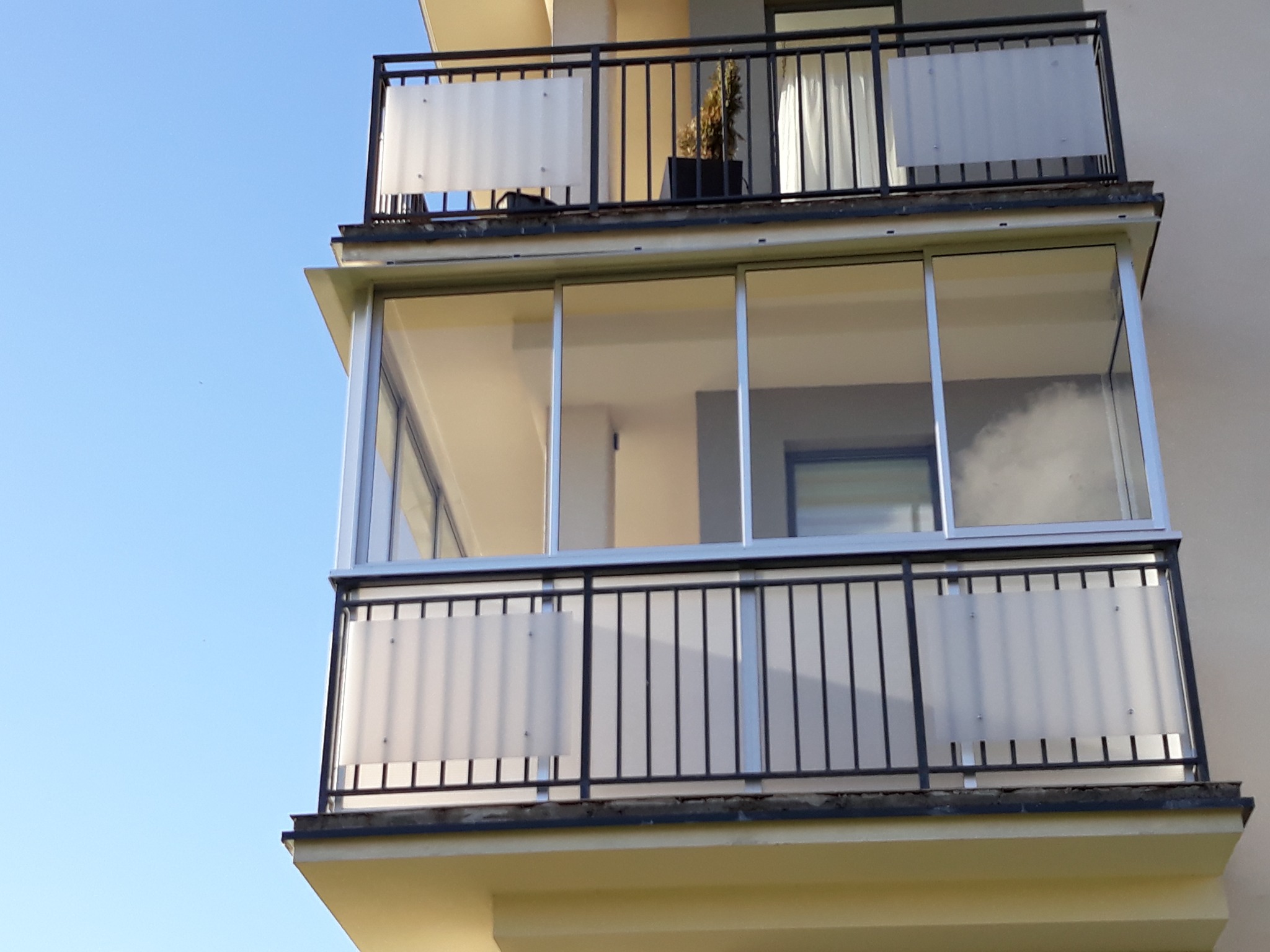 Zabudowany balkon z metalową balustradą i mlecznym szkłem, widok z dołu na dwa piętra budynku.