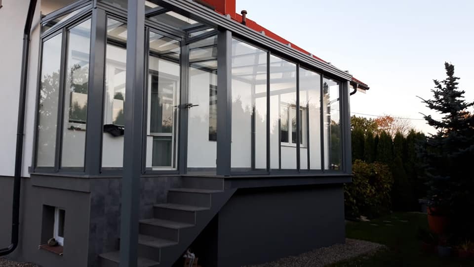 Szklana konstrukcja tarasowa z metalową ramą, schody prowadzące do wejścia, widok z zewnątrz, szara kolorystyka, widoczny fragment ogrodu.