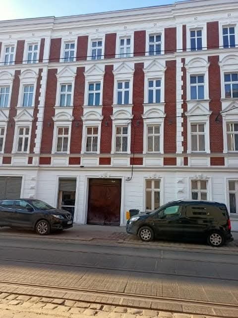 Odnowiona fasada kamienicy z czerwono-białymi zdobieniami, widoczna z poziomu ulicy z zaparkowanymi samochodami. Klasyczna architektura miejska po renowacji.