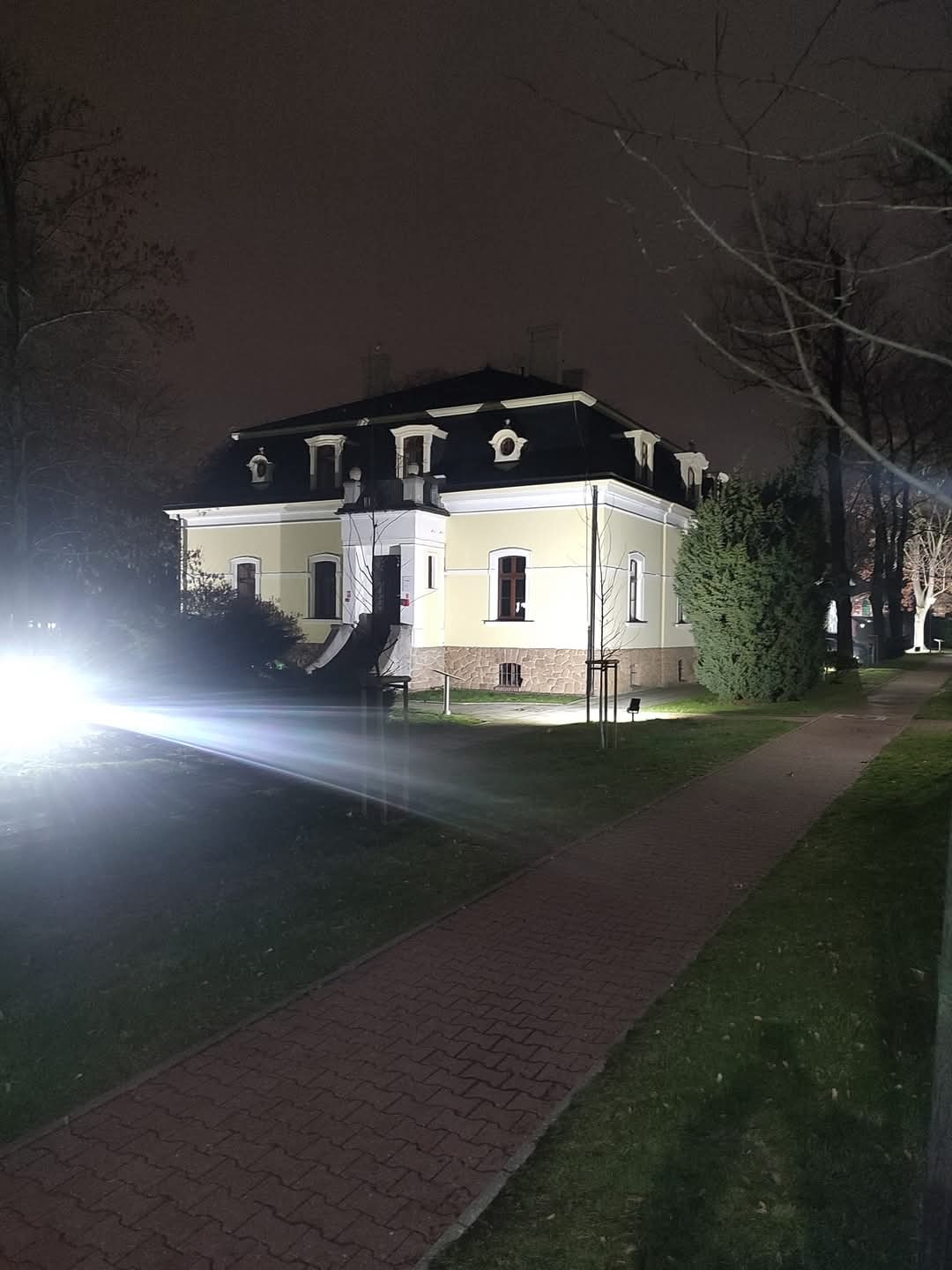 Oświetlony nocą, odnowiony budynek z żółtą elewacją, czarnym dachem i białymi detalami architektonicznymi. Widoczny chodnik z kostki i drzewa bez liści.