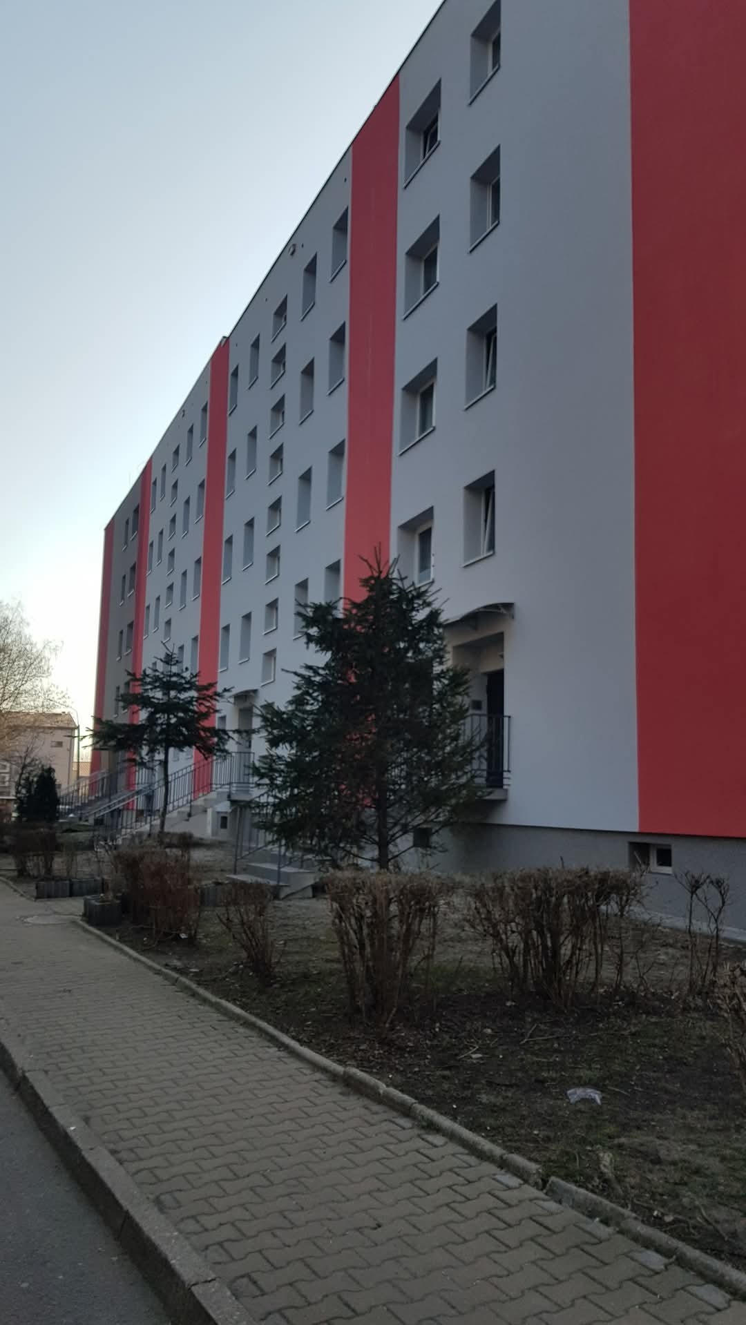 Wielopiętrowy budynek po renowacji elewacji, z szarymi ścianami i czerwonymi pasami, widoczny z poziomu chodnika. Uwagę zwracają małe drzewka przed budynkiem.