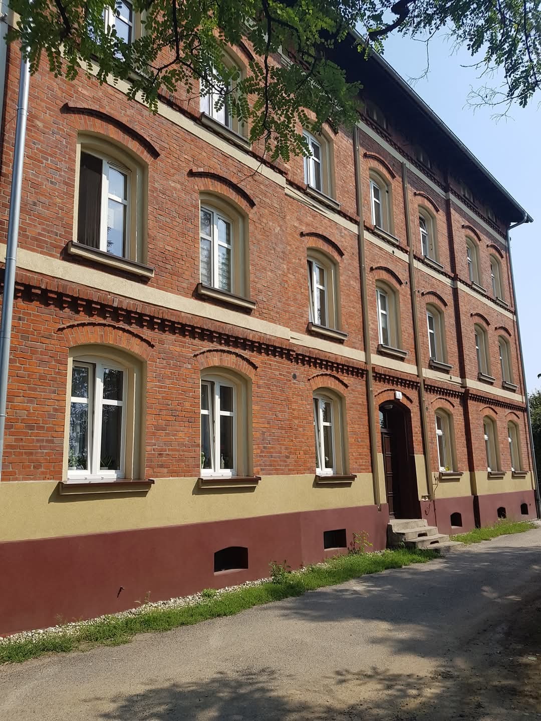 Dwupiętrowy budynek z czerwonej cegły z jasnymi oknami i ciemnym pasem u dołu elewacji, widoczny z perspektywy ulicy w słoneczny dzień.