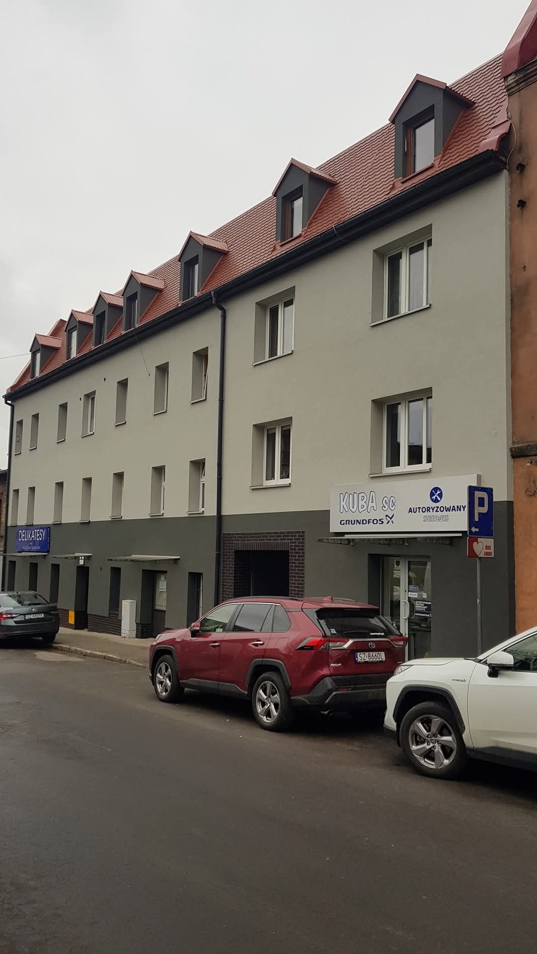 Odnowiona elewacja kamienicy z rzędem okien dachowych, pomalowana na szaro z ciemniejszym pasem u dołu. Widoczne zaparkowane samochody i szyld sklepu.