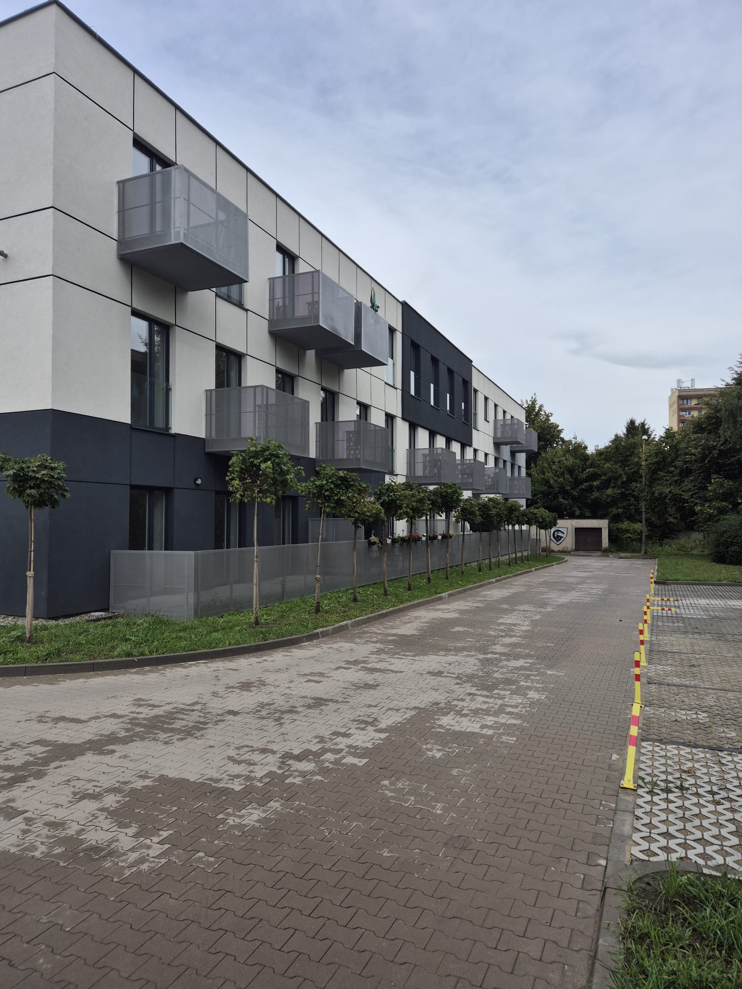 Nowoczesny budynek wielorodzinny z szaro-białą elewacją i metalowymi balkonami, otoczony rzędem małych drzew i brukowanym parkingiem w pochmurny dzień.