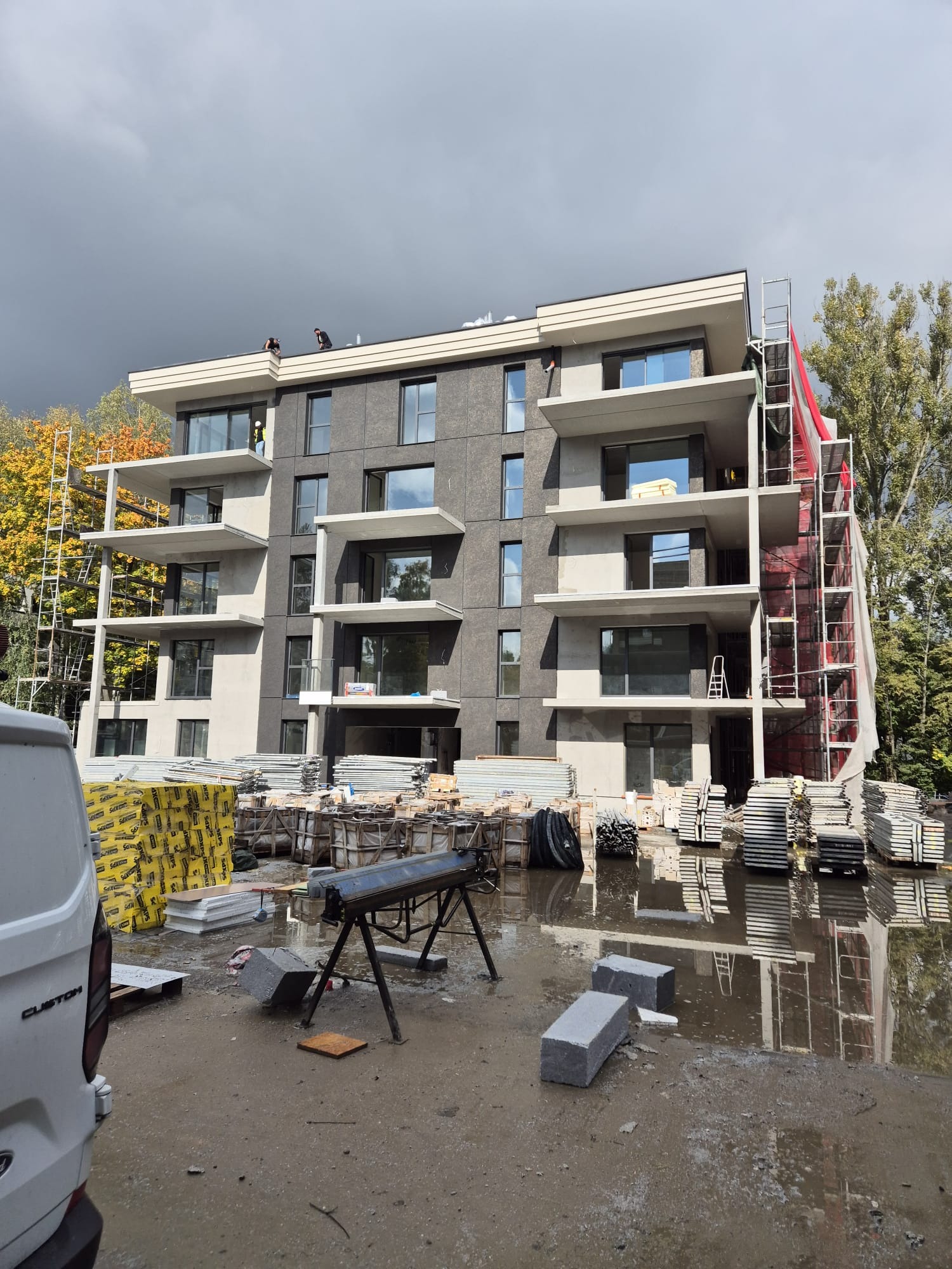 Wielopiętrowy budynek w trakcie prac elewacyjnych, widoczne rusztowania, materiały budowlane i pracownicy na dachu. Pochmurne niebo w tle.