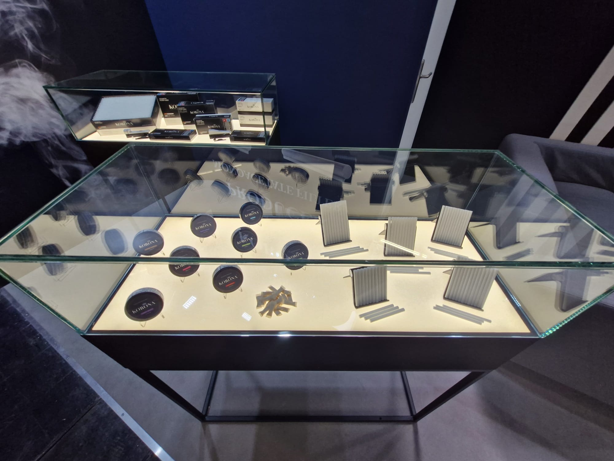 Szklana gablota z ekspozycją produktów marki Korona: pudełka, puszki i akcesoria do palenia na podświetlonym blacie. U góry dodatkowa półka z podobnymi produktami, tło w odcieniach czerni i granatu.