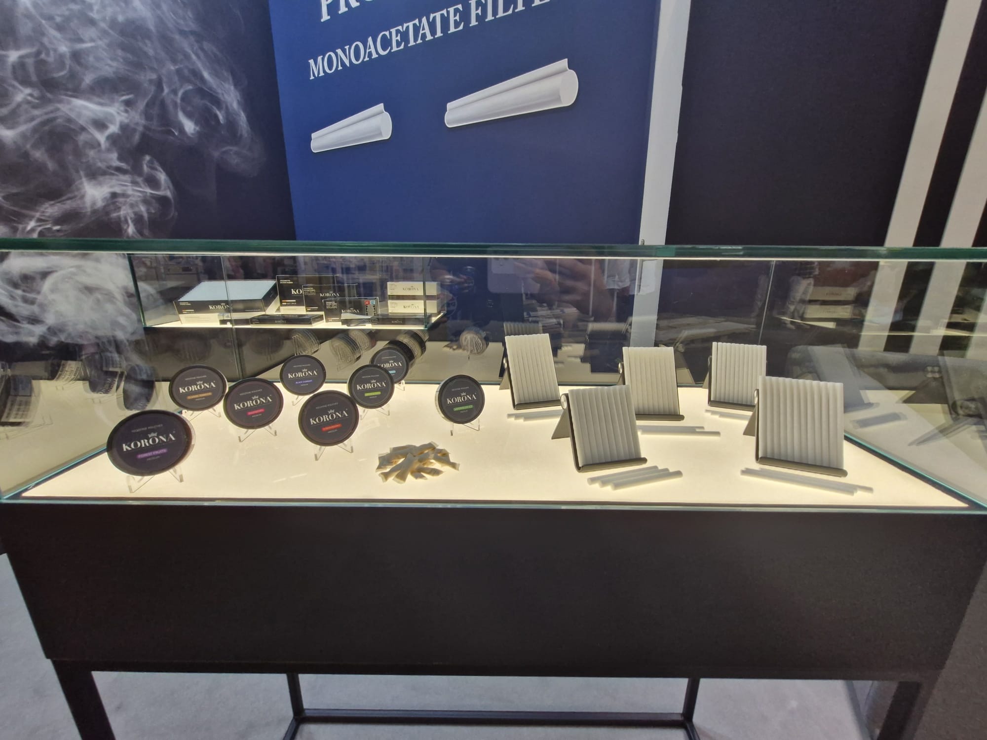 Ekspozycja w szklanej gablocie z produktami marki Korona: okrągłe puszki z tytoniem, filtry monoacetatowe i papierosy.