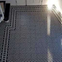 Czarno-biała mozaika podłogowa z geometrycznym wzorem trójkątów, ułożona w korytarzu przy schodach, z widocznym fragmentem okna wpadającego światła.