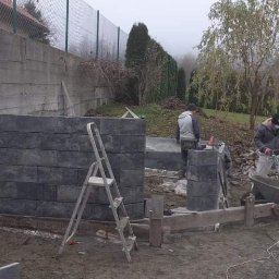 Remonty i Wykończenia Adam Światłoń - Budowa murku oporowego z szarych bloczków betonowych, widoczni dwaj pracownicy w trakcie pracy, drabina oparta o konstrukcję, tło stanowią drzewa i skarpa.