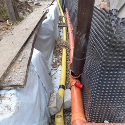 Remonty i Wykończenia Adam Światłoń - Wykop z izolacją pionową fundamentu, drenaż z rurami w kolorach żółtym i pomarańczowym, podsypka żwirowa, folia izolacyjna, widoczna ściana fundamentowa zabezpieczona folią kubełkową.