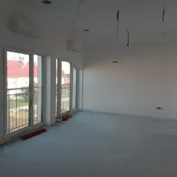 Wnętrze w stanie deweloperskim po malowaniu ścian na biało, z widocznymi oknami balkonowymi, klimatyzatorami, wystającymi przewodami z sufitu i przygotowaną podłogą.