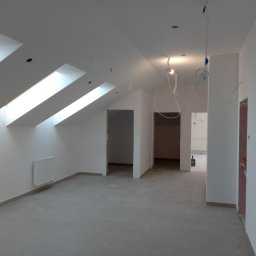 Remonty i Wykończenia Adam Światłoń - Poddasze po remoncie z trzema oknami dachowymi, grzejnikiem na ścianie, szarymi płytkami na podłodze i przewodami elektrycznymi zwisającymi z sufitu.