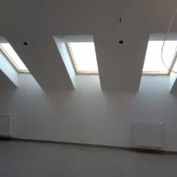 Poddasze z trzema oknami dachowymi w drewnianych ramach, świeżo pomalowane ściany na biało, dwa białe grzejniki, widoczne kable wystające z sufitu.