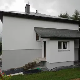 Dwukondygnacyjny dom z szarą elewacją, widoczne schody wejściowe, metalowy dach i przygotowane otoczenie z folią ochronną i kwiatami w doniczce.