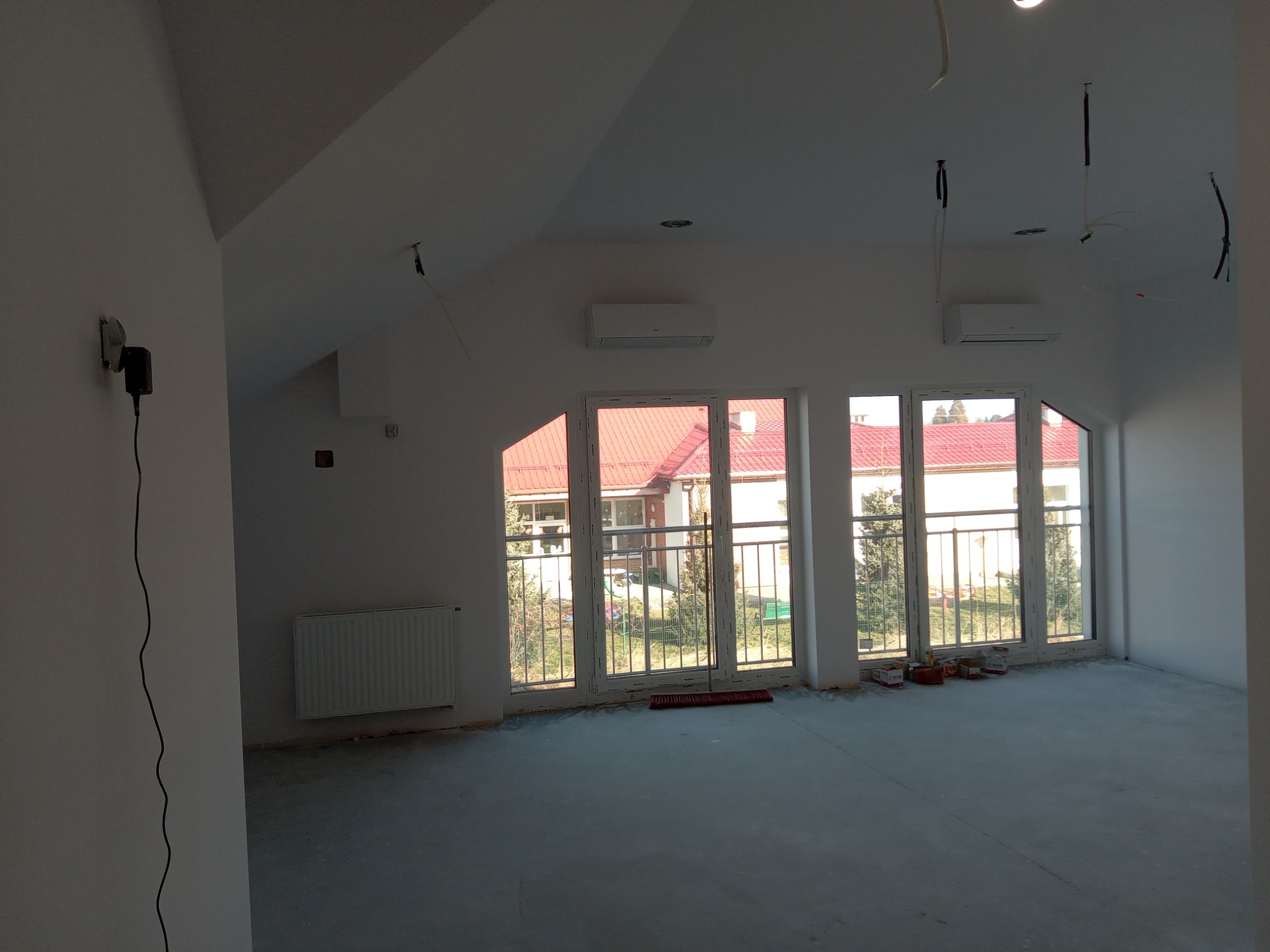 Poddasze w trakcie prac wykończeniowych: ściany pomalowane na biało, widoczne okna z widokiem na zewnątrz, grzejnik, zainstalowana klimatyzacja, wystające kable z sufitu, surowa podłoga.
