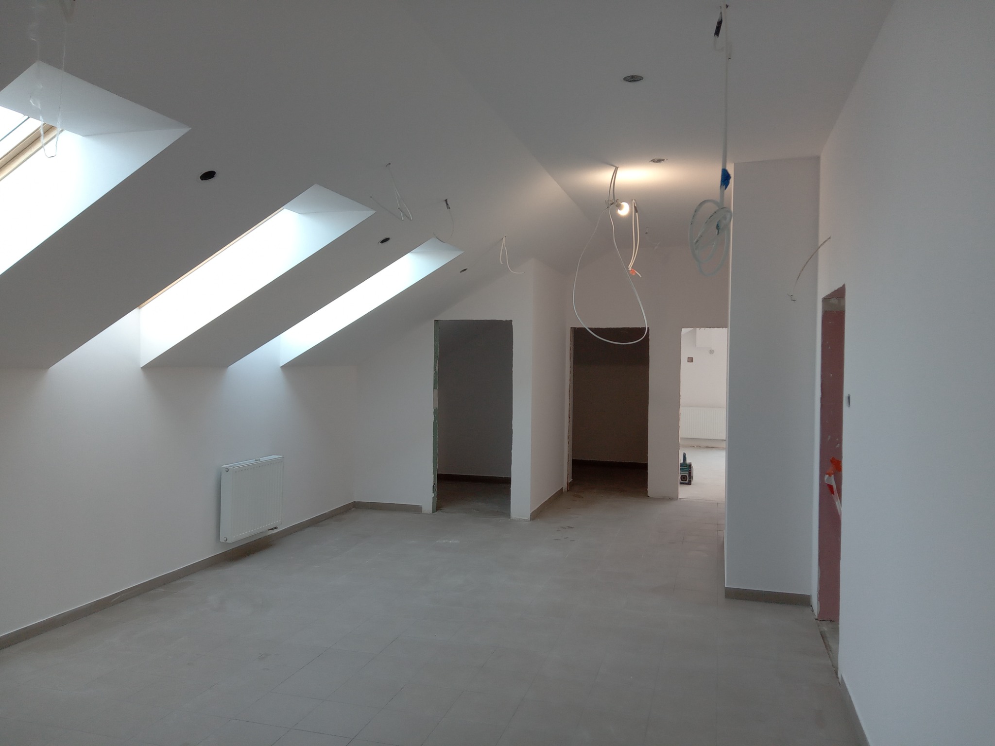 Poddasze po remoncie z trzema oknami dachowymi, grzejnikiem na ścianie, szarymi płytkami na podłodze i przewodami elektrycznymi zwisającymi z sufitu.