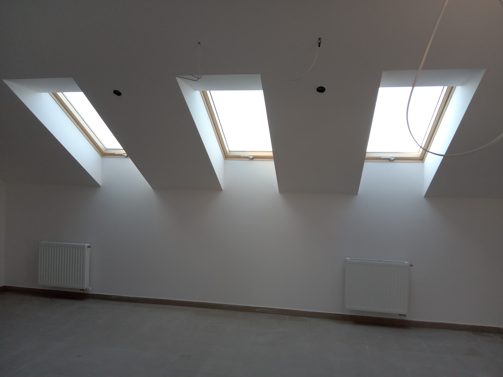 Poddasze z trzema oknami dachowymi w drewnianych ramach, świeżo pomalowane ściany na biało, dwa białe grzejniki, widoczne kable wystające z sufitu.