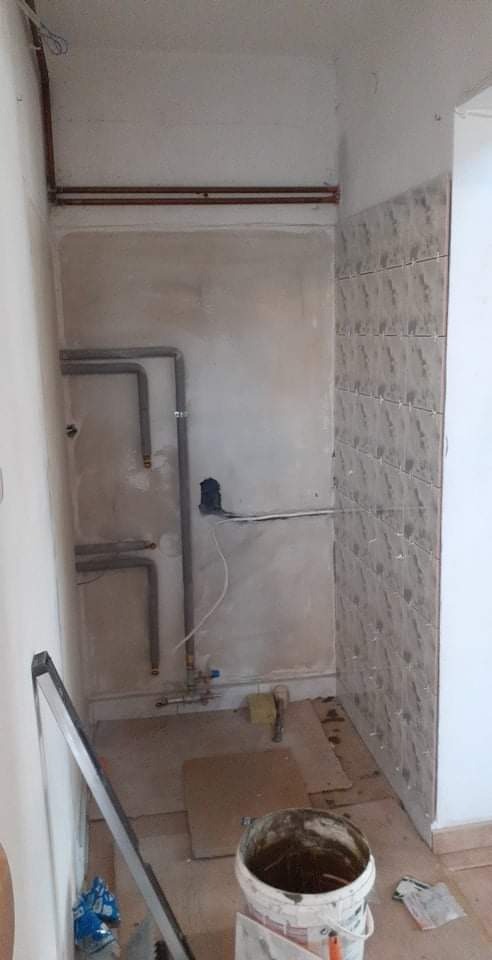 Wnętrze łazienki w trakcie remontu: częściowo ułożone płytki na ścianie, widoczne rury, puszka elektryczna, poziomica oparta o ścianę, wiadro z zaprawą i fragmenty podłogi zabezpieczonej kartonem.