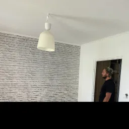 Pokój z białą ceglaną ścianą, wiszącą lampą z abażurem i mężczyzną w czarnej koszulce i czapce z daszkiem w tle, stojącym w otwartych drzwiach.