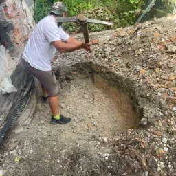 Mężczyzna w czapce z daszkiem kopie dół w ziemi kilofem przy fundamentach budynku, widoczna izolacja fundamentów.