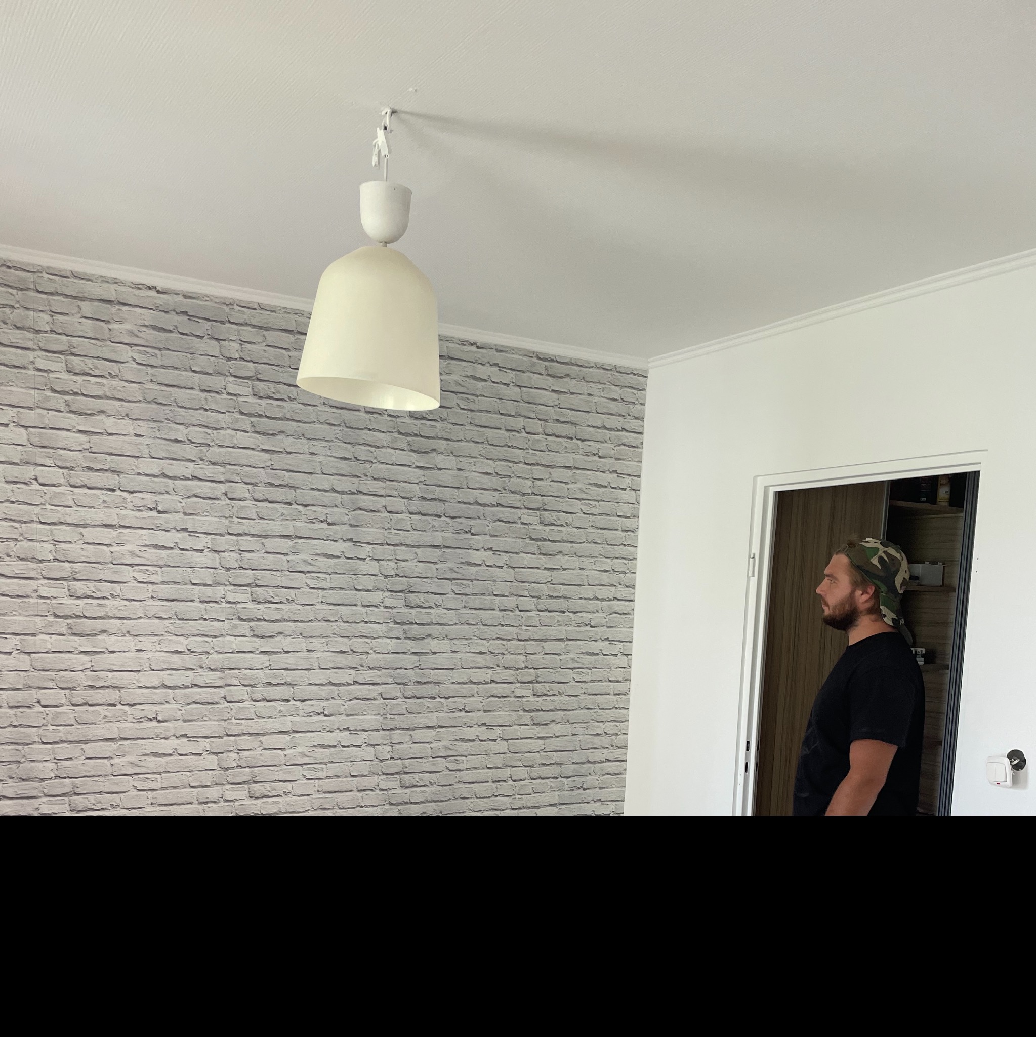 Pokój z białą ceglaną ścianą, wiszącą lampą z abażurem i mężczyzną w czarnej koszulce i czapce z daszkiem w tle, stojącym w otwartych drzwiach.