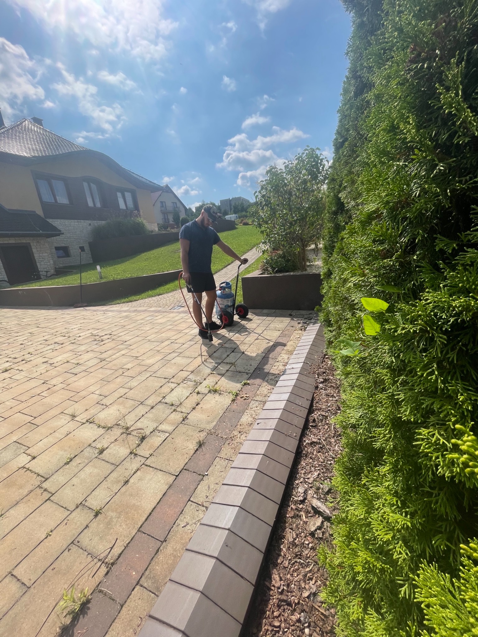 Mężczyzna czyści kostkę brukową na podjeździe myjką ciśnieniową, widoczny dom w tle i żywopłot z boku.